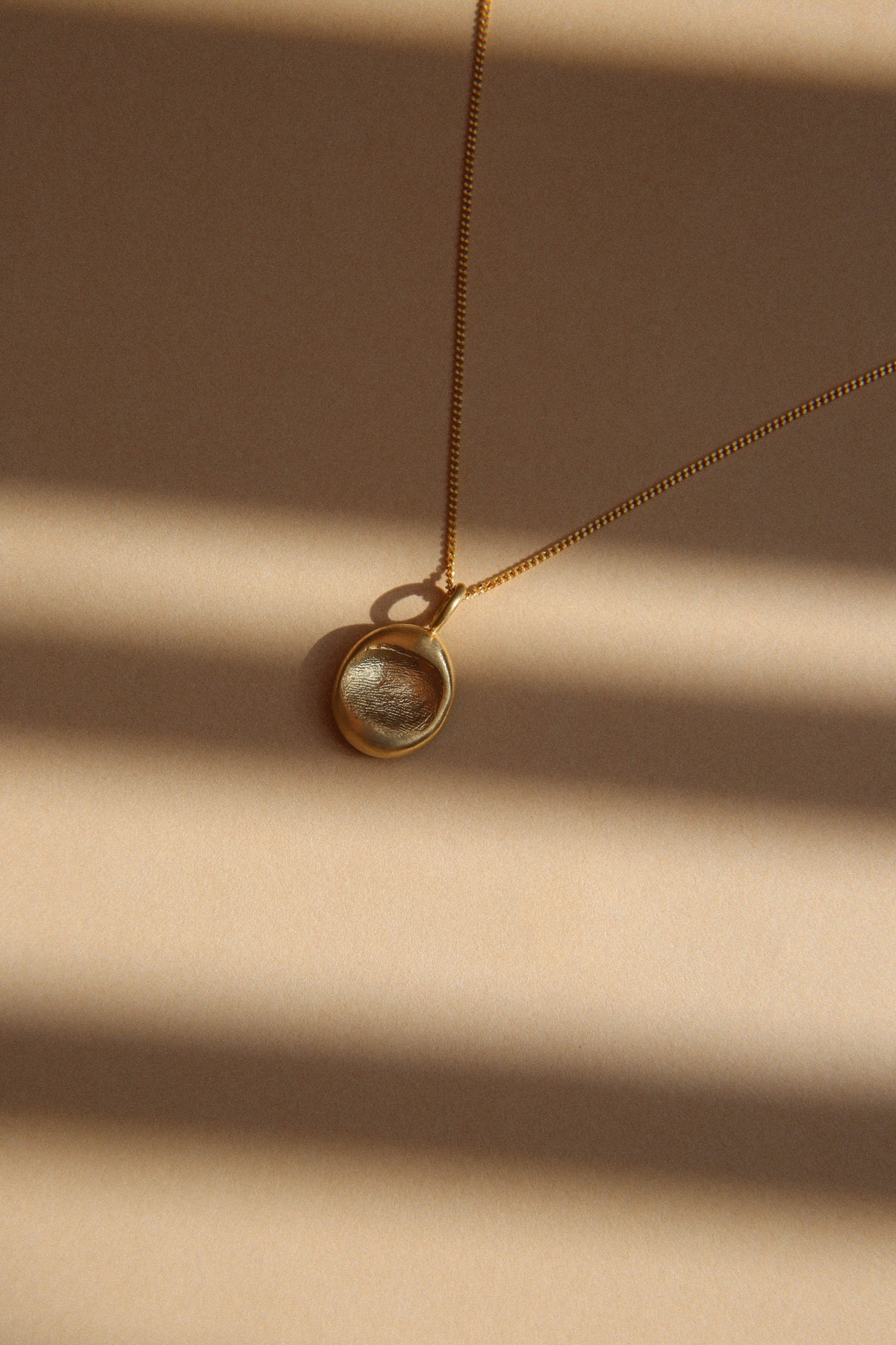 The details: small oval gold fingerprint pendant on a fine chain, in stripes of sunlight