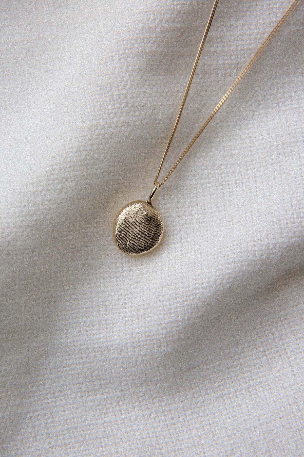 The details: gold fingerprint pendant on a fine chain, laid on white fabric